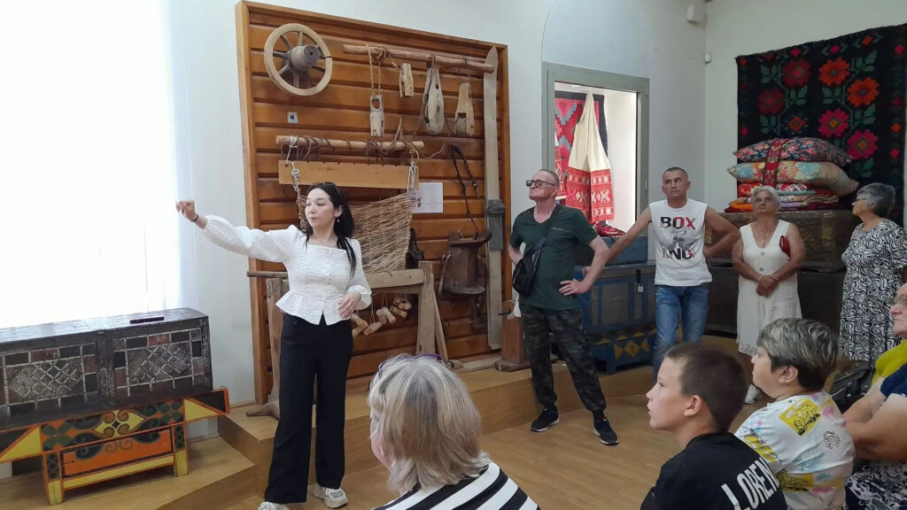 Экскурсовод рассказывает участникам об экспозиции, посвящённой традиционному быту и ремёслам Башкортостана. За её спиной представлены старинные предметы домашнего обихода и ткацкий станок.