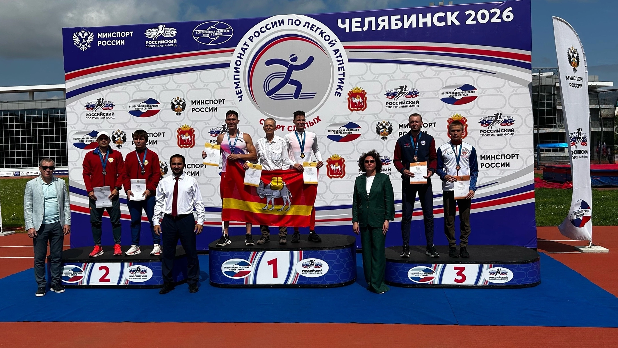 Пьедестал чемпионата России по лёгкой атлетике в Челябинске 2026 года: на трёх ступенях стоят призёры с медалями и грамотами, в центре победители держат красно‑жёлтый флаг с верблюдом, позади большой баннер Минспорта России.