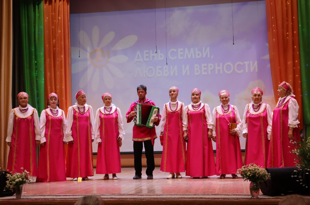 На сцене ансамбль: девять пожилых женщин в одинаковых ярко‑розовых русских сарафанах и кокошниках, в центре мужчина в розовой рубахе играет на аккордеоне