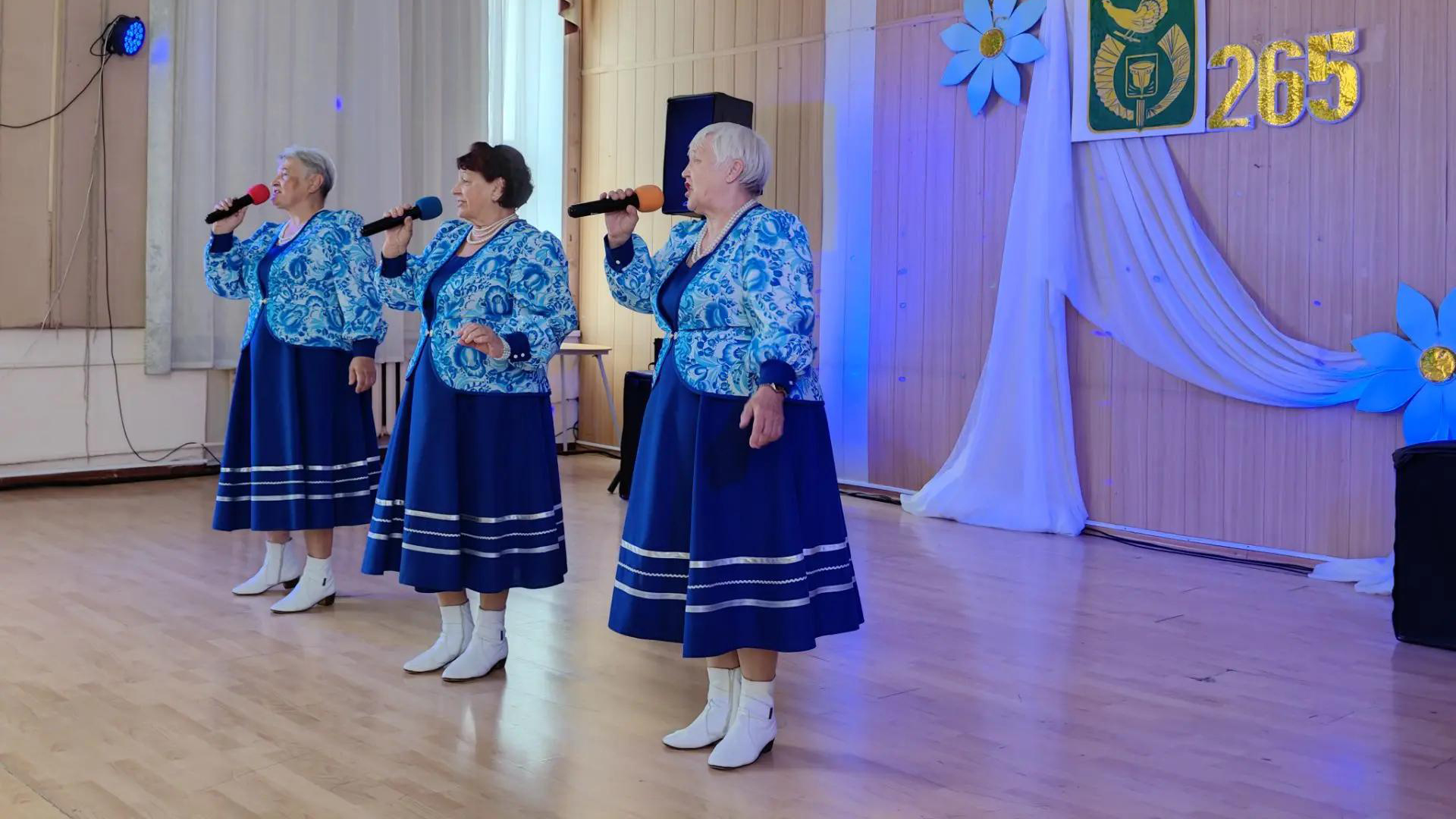 Три пожилые женщины в одинаковых синих костюмах поют на сцене в микрофоны, на заднем плане украшения и золотая цифра «265».