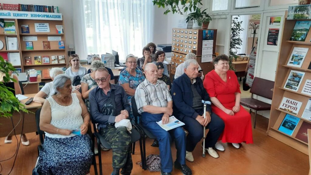 Группа людей сидит на стульях в библиотеке на мероприятии, вокруг книжные стеллажи и стенд с надписью «НАШ ГЕРОЙ».