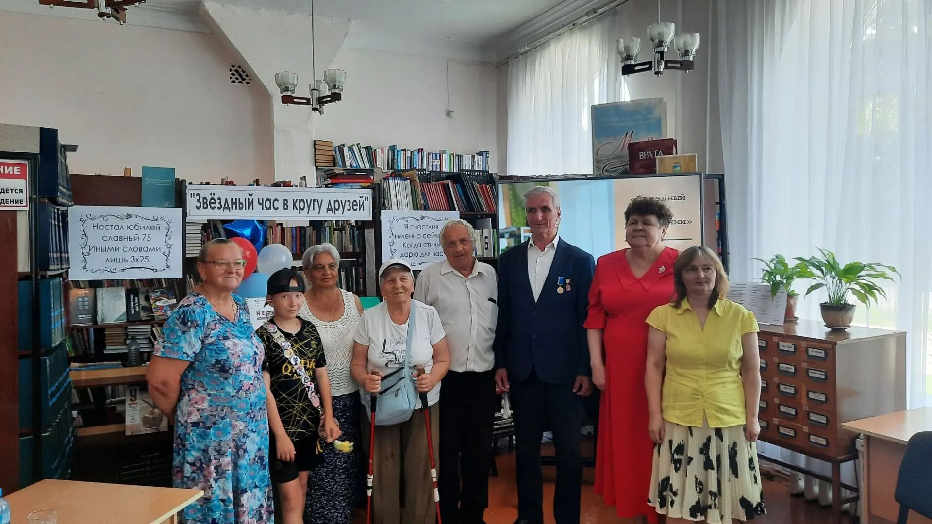 Группа из восьми человек стоит в ряд в библиотеке на фоне книжных стеллажей и плакатов с надписями «Звёздный час в кругу друзей»