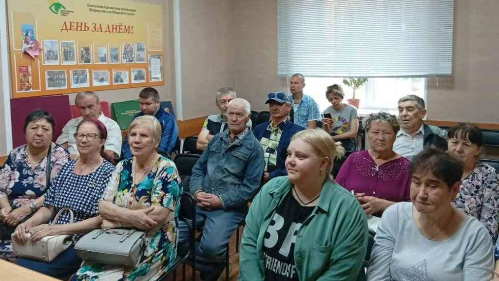 группа людей на встрече. На стене слева большой жёлтый стенд с надписью «ДЕНЬ ЗА ДНЁМ!» и фотографиями. Люди сидят плотными рядами и слушают кого-то перед ними, вне кадра.