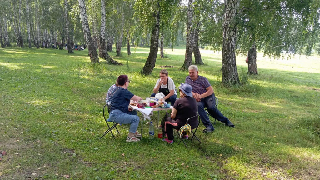 Пятеро человек сидят за маленьким столом на стульчиках в берёзовой роще и устраивают пикник, на столе еда и напитки.