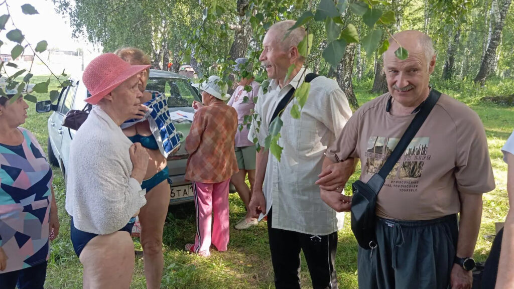 Группа людей общается в тени деревьев у припаркованной машины, часть в купальниках, двое мужчин справа улыбаются и держатся под руку.