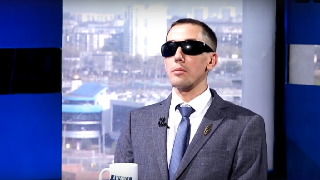 Александр Рожков в деловом костюме и тёмно-синим галстуком, в больших чёрных солнцезащитных очках, сидит в студии на фоне размытых городских зданий.