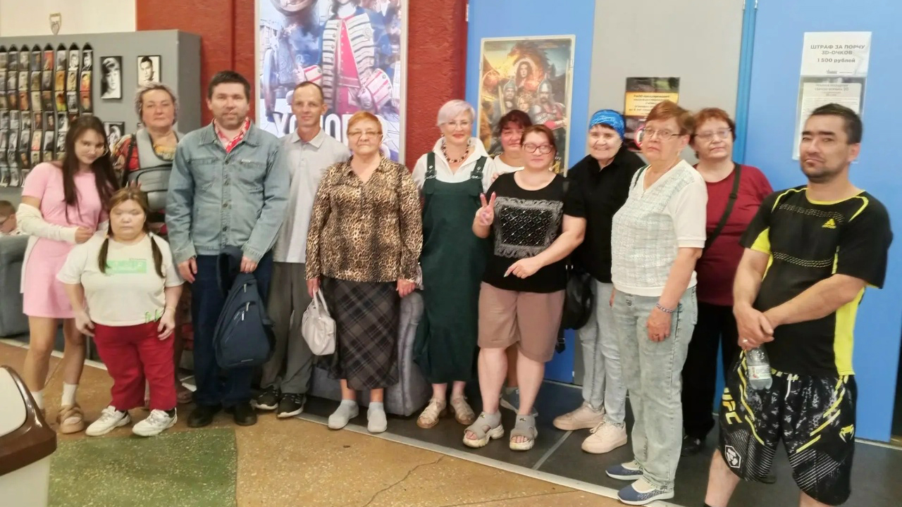 На фото большая группа людей разных возрастов стоит в помещении у входа в кинотеатр: за ними цветные постеры фильмов и таблички на стене,