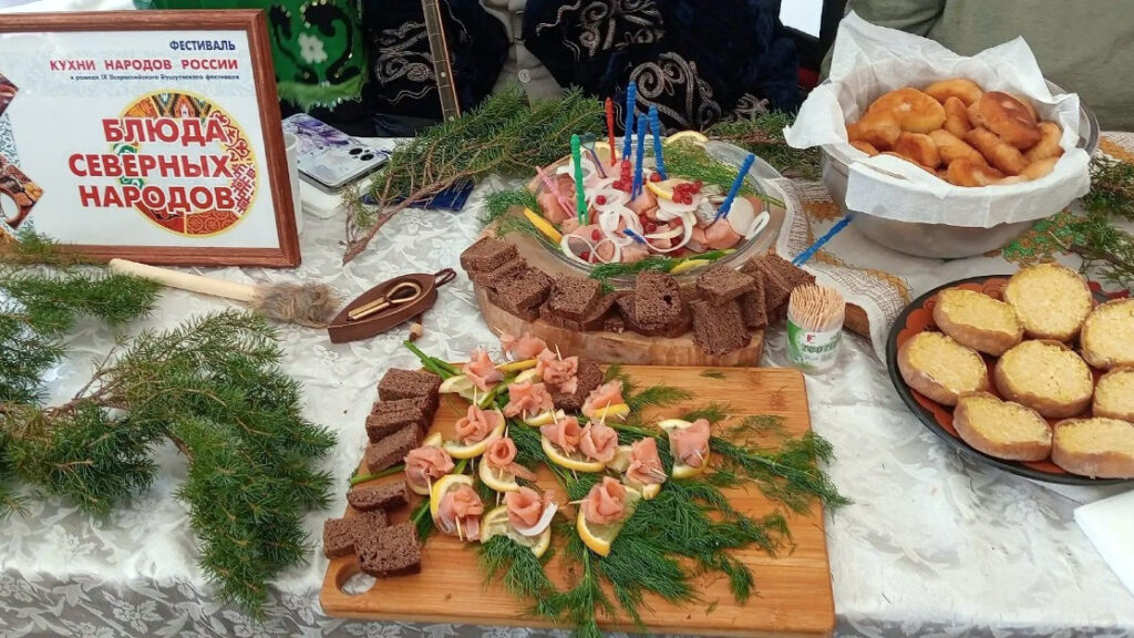 На столе представлены блюда северных народов: бутерброды с рыбой, хлеб, выпечка и закуски, украшенные зеленью.