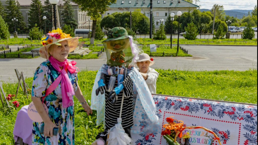 Участница фестиваля стоят у конкурсной площадки рядом с необычным огородным пугалом в шляпе, перчатках и прозрачной накидке