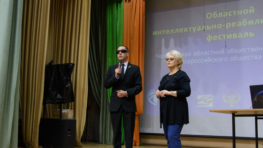 Мужчина в тёмном костюме выступает с микрофоном на сцене; рядом стоит женщина, на экране — заставка областного интеллектуально-реабилитационного фестиваля.