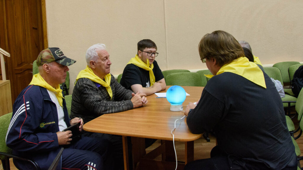 Команда в жёлтых косынках обсуждает задание за столом; в центре установлен светящийся игровой сигнал.