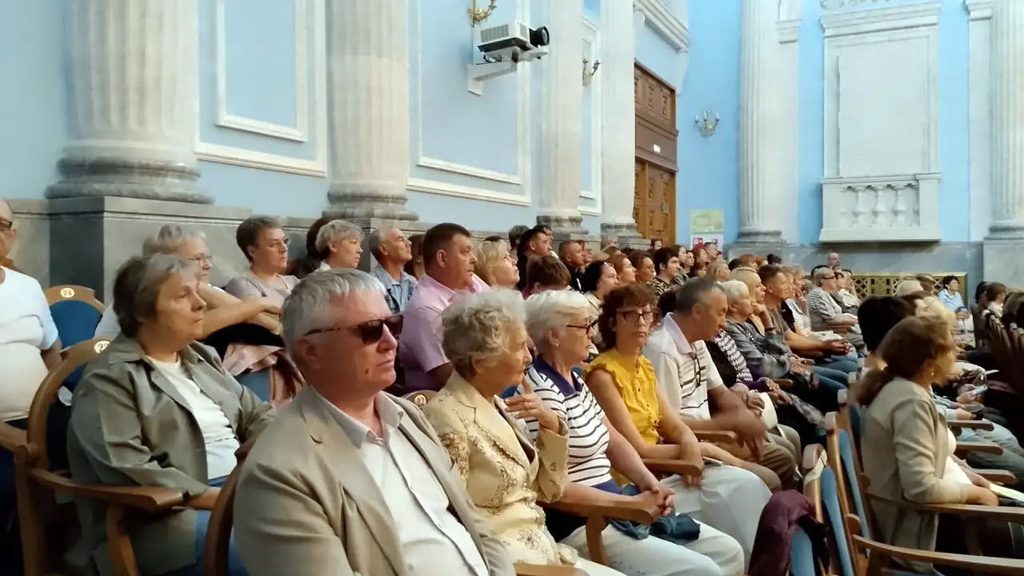Зрители сидят в зале во время концерта органной музыки; среди них есть люди в темных очках.
