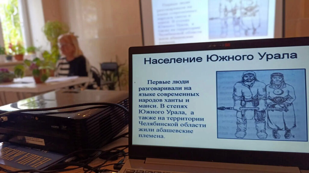 На экране ноутбука показан слайд «Население Южного Урала» с иллюстрацией древних жителей; на заднем плане видна участница встречи