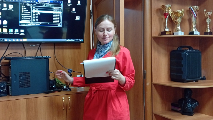 Женщина в красном платье читает текст по листам; за ней видны экран компьютера и полки с кубками.