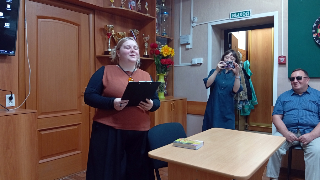 Женщина читает текст перед участниками встречи; рядом другая участница фотографирует выступление на телефон.