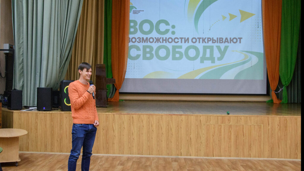 Александр Новичихин с микрофоном выступает перед участниками встречи на фоне экрана с названием «ВОС: возможности открывают свободу».