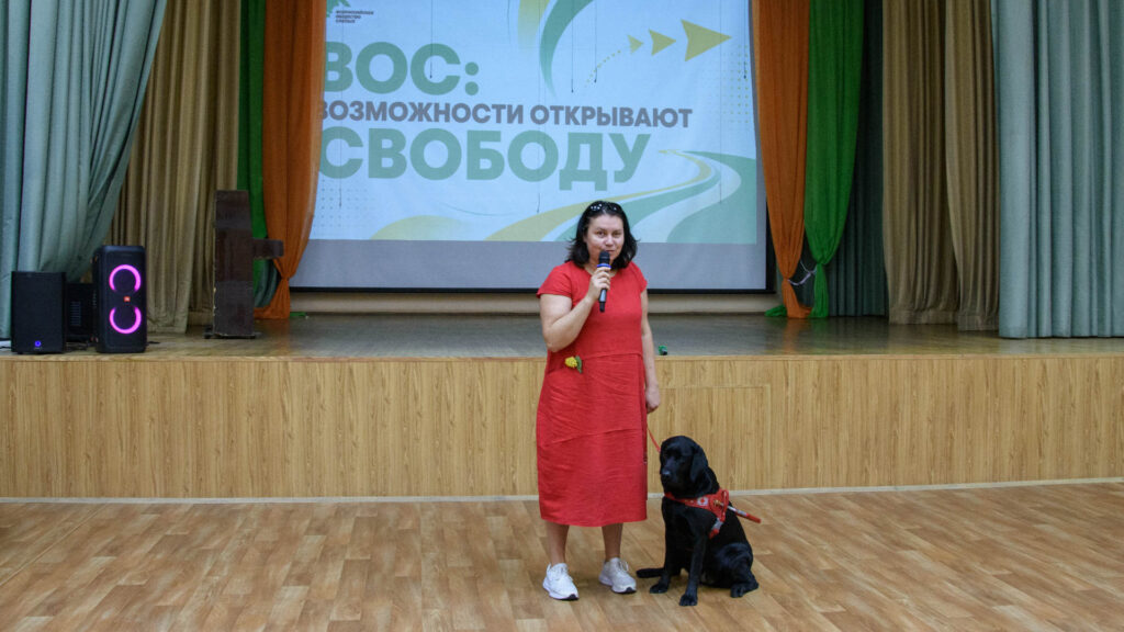 Надежда Завей-Борода с микрофоном рассказывает о собаках-проводниках; рядом с ней сидит чёрный лабрадор в специальной шлейке.