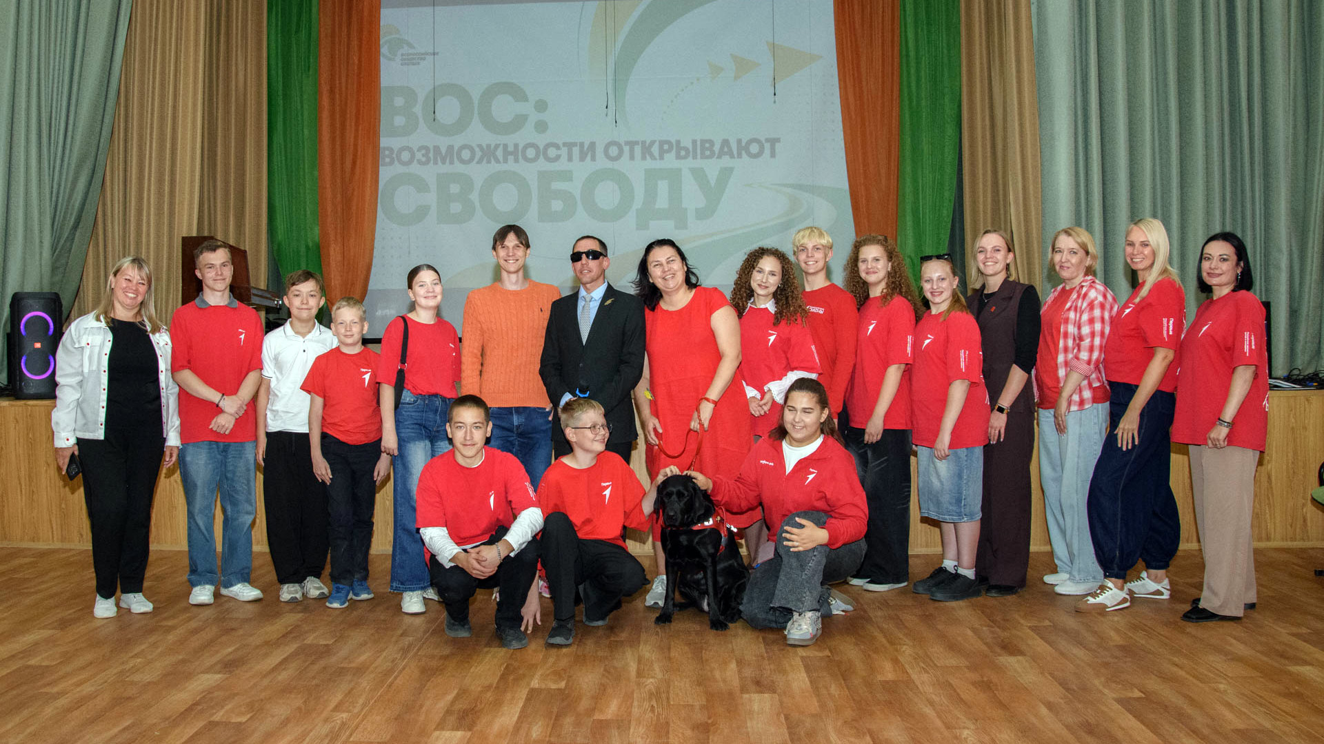 Участники и организаторы встречи «ВОС: возможности открывают свободу» фотографируются вместе на сцене; в центре сидит чёрная собака-проводник.