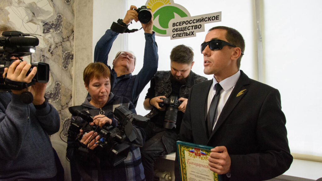 Председатель ЧООО ВОС Александр Рожков вручает благодарственное письмо Олегу Герберу; важный момент одновременно снимают несколько журналистов, телеоператоров и фотографов.