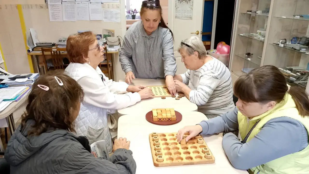 Участники Чебаркульской МО ВОС за столом осваивают адаптированные настольные игры; инструктор Светлана Кушлакова помогает разобраться с правилами и ходами.