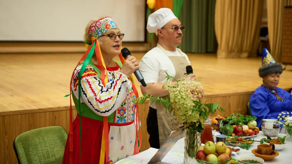 Участники конкурса национальных блюд представляют белорусскую кухню; женщина в народном костюме выступает с микрофоном, рядом мужчина в поварском колпаке, на столе — блюда и угощения.