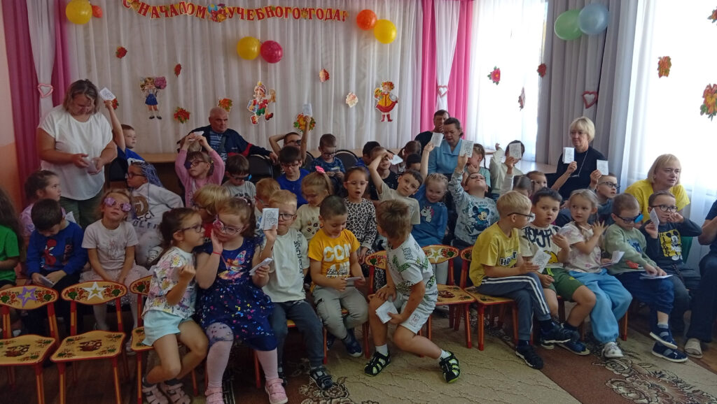 Воспитанники детского сада сидят в зале после представления и показывают сделанные своими руками книжки-раскладушки.