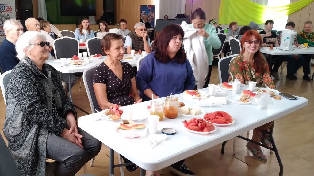 Участники праздника сидят за столами с угощениями, домашними заготовками, арбузом и дыней.