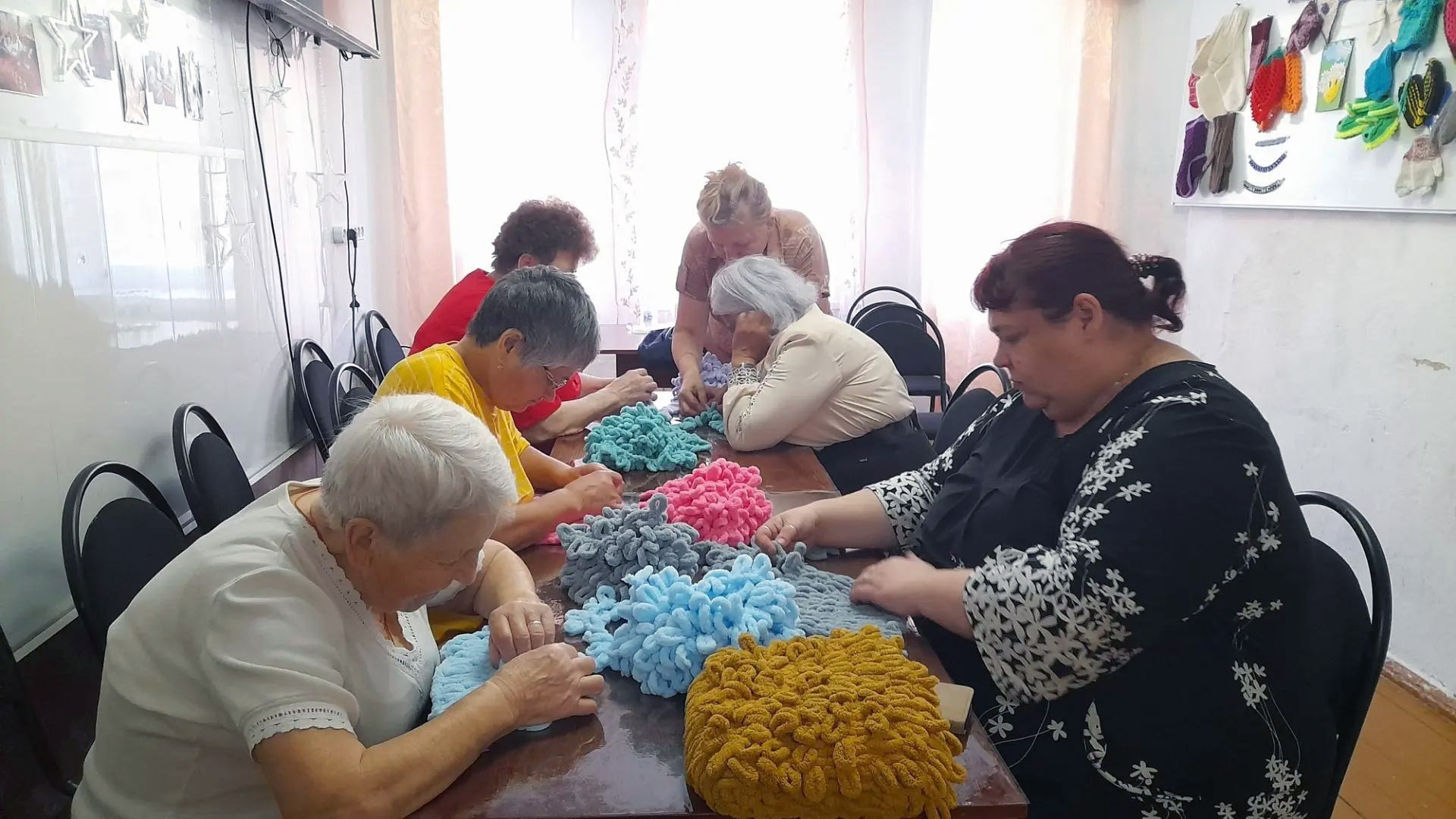 Женщины сосредоточенно работают над вязаными подушками-думками, специалист помогает им.