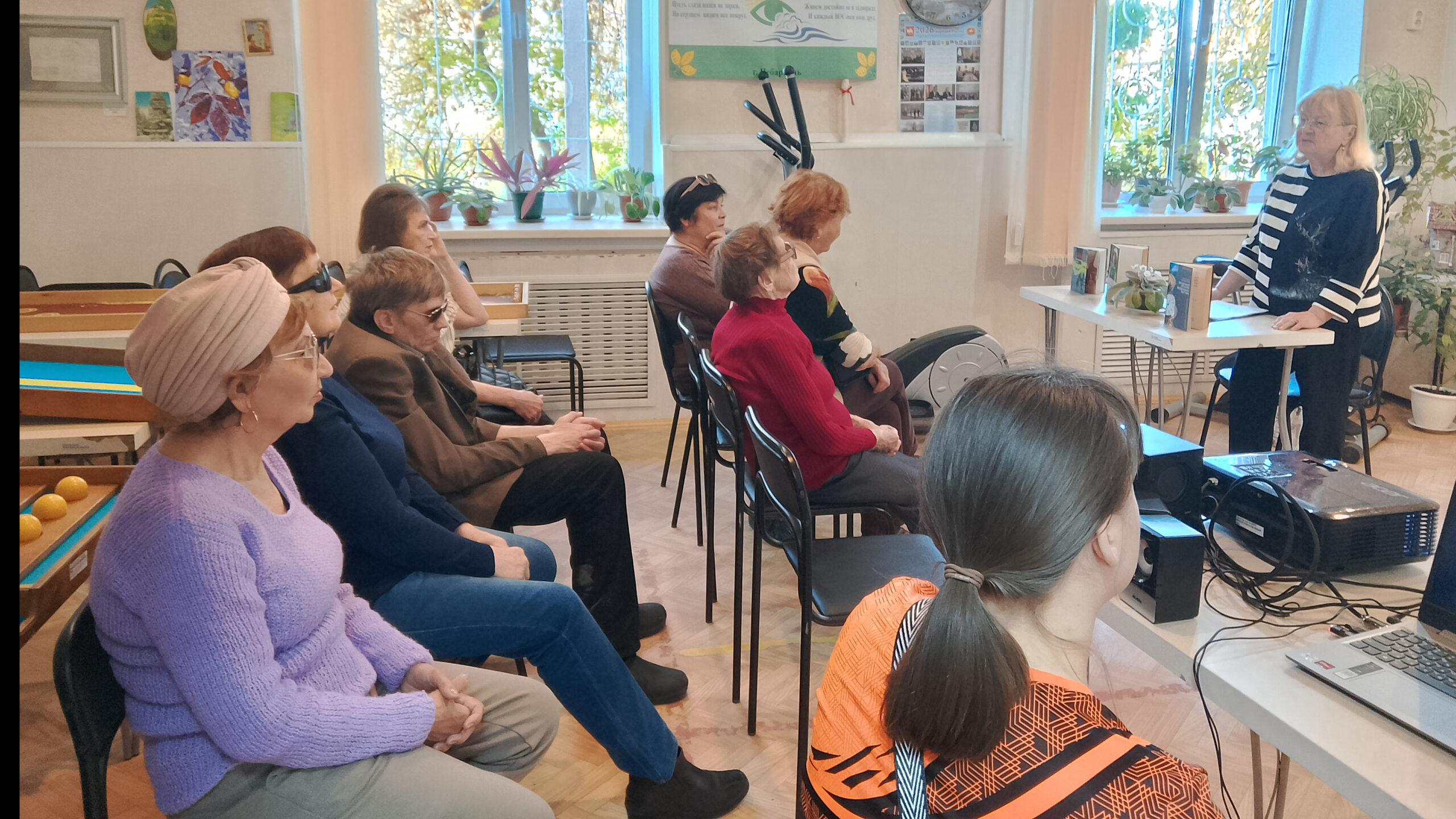 Женщина стоит у стола и рассказывает о книгах группе слушателей, сидящих на стульях в помещении. На столе перед ней выставлены книги, рядом находятся компьютер и проектор.