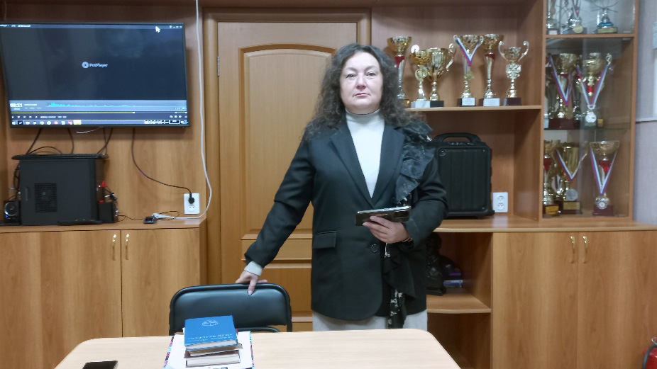 женщина в классическом черном пиджаке и светлой водолазке, стоящая в официальном кабинете с кубками и офисной техникой