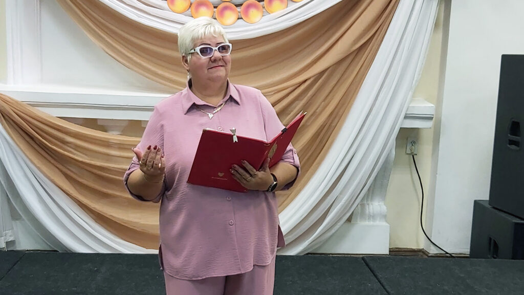 Женщина в розовом костюме держит в руках красную папку для выступлений.