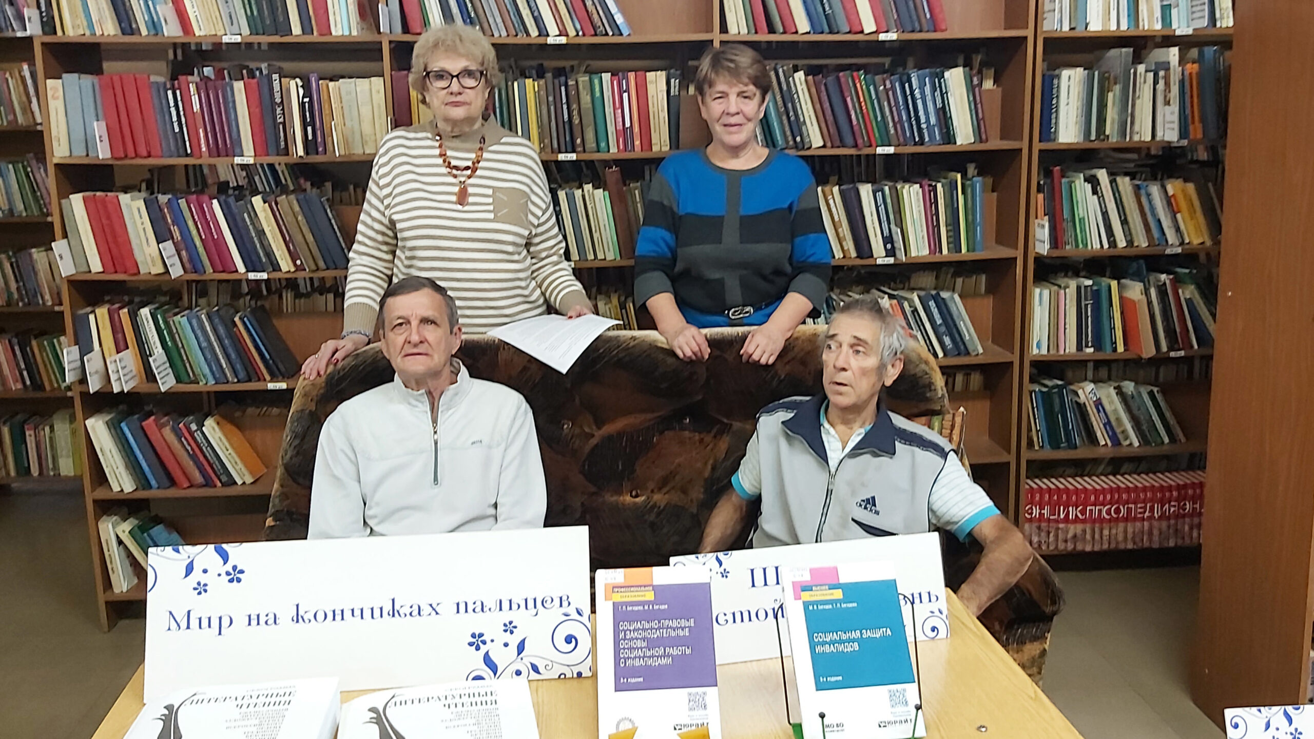 Четыре человека позируют на фоне книжных стеллажей в библиотеке. Перед ними на столе лежат книги, изданные шрифтом Брайля, и стоят учебные пособия по социальной работе и защите прав инвалидов.