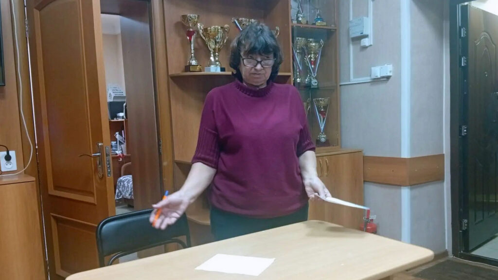женщина средних лет в бордовом свитере и очках, стоящая за светлой партой в светлом помещении с деревянной мебелью и кубками на полках
