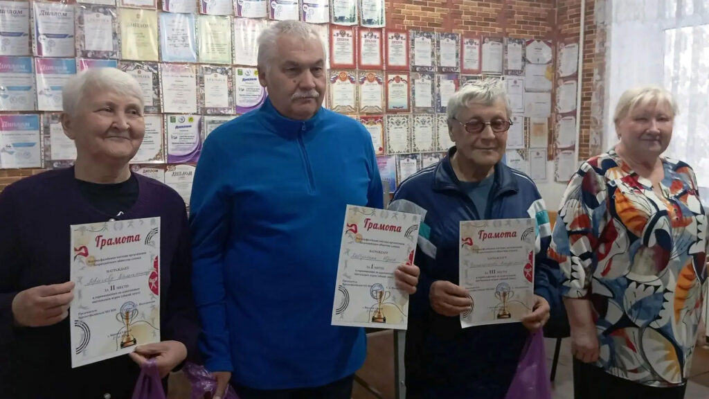 Три участника чемпионата по адаптивным настольным играм стоят с грамотами за призовые места. Рядом с ними — организатор соревнований. На заднем плане размещены дипломы и грамоты.