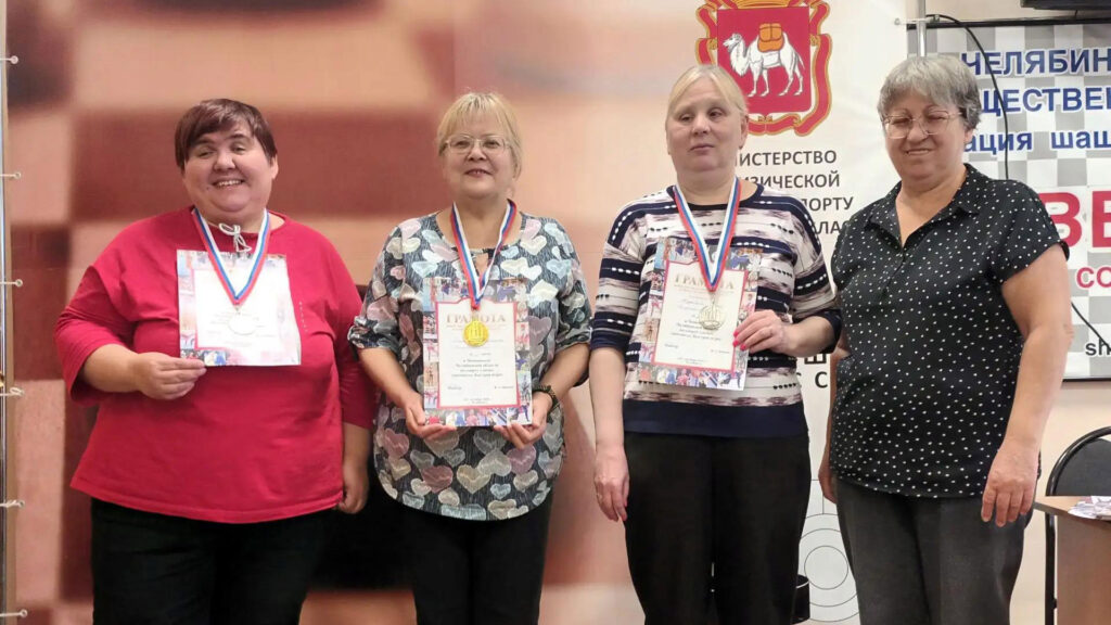 Три женщины — призёры чемпионата Челябинской области по шахматам среди спортсменов с нарушением зрения — стоят с медалями и грамотами. Рядом с ними находится представительница организаторов соревнований.