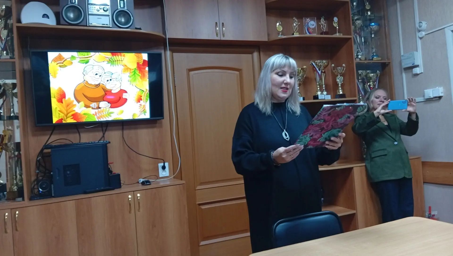 На снимке запечатлена женщина в чёрной удлинённой тунике или платье свободного силуэта, выступающая с документом в руках.